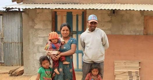 A family of five standing outside their completed house, the parents holding two small children and a third standing at their side