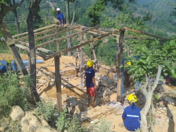 Workers in yellow hard hats raising a timber frame on a dry stone plinth on a cleared hillside plot